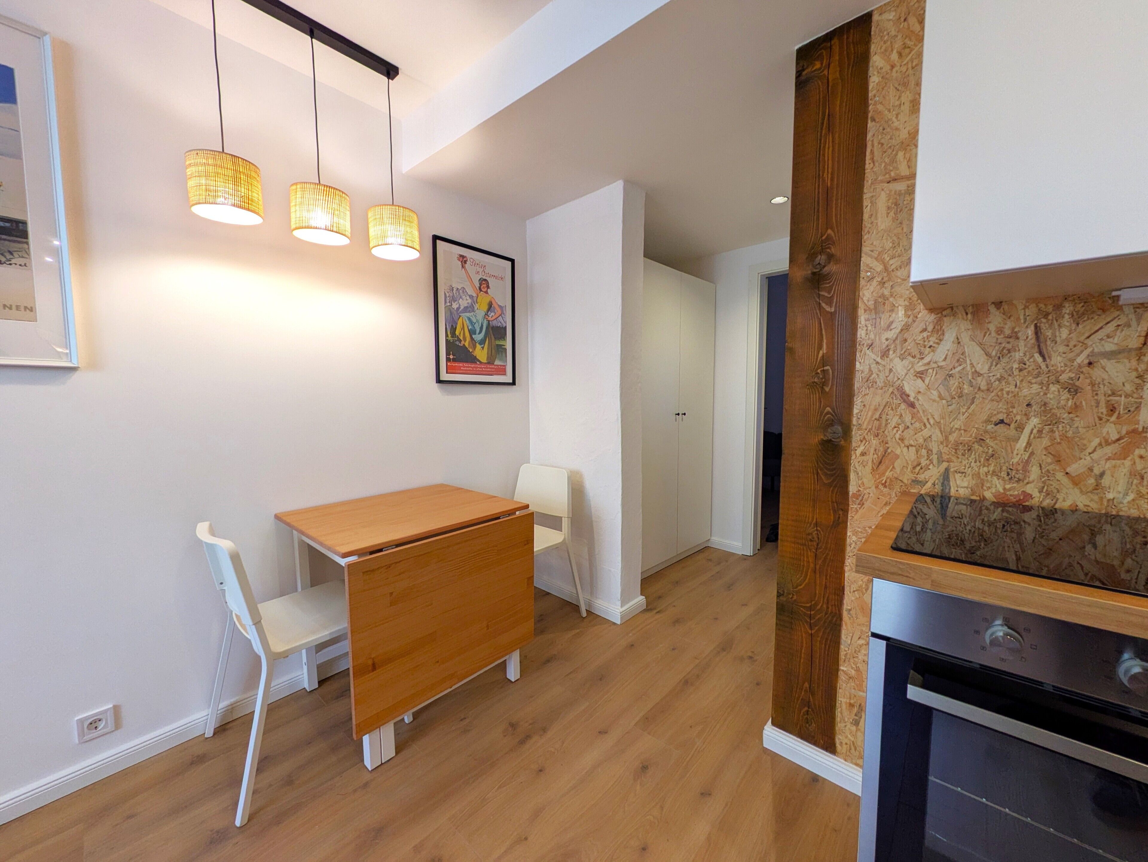 Una cocina moderna con horno empotrado y un mueble de madera. Hay un comedor con mesa de madera y sillas blancas.