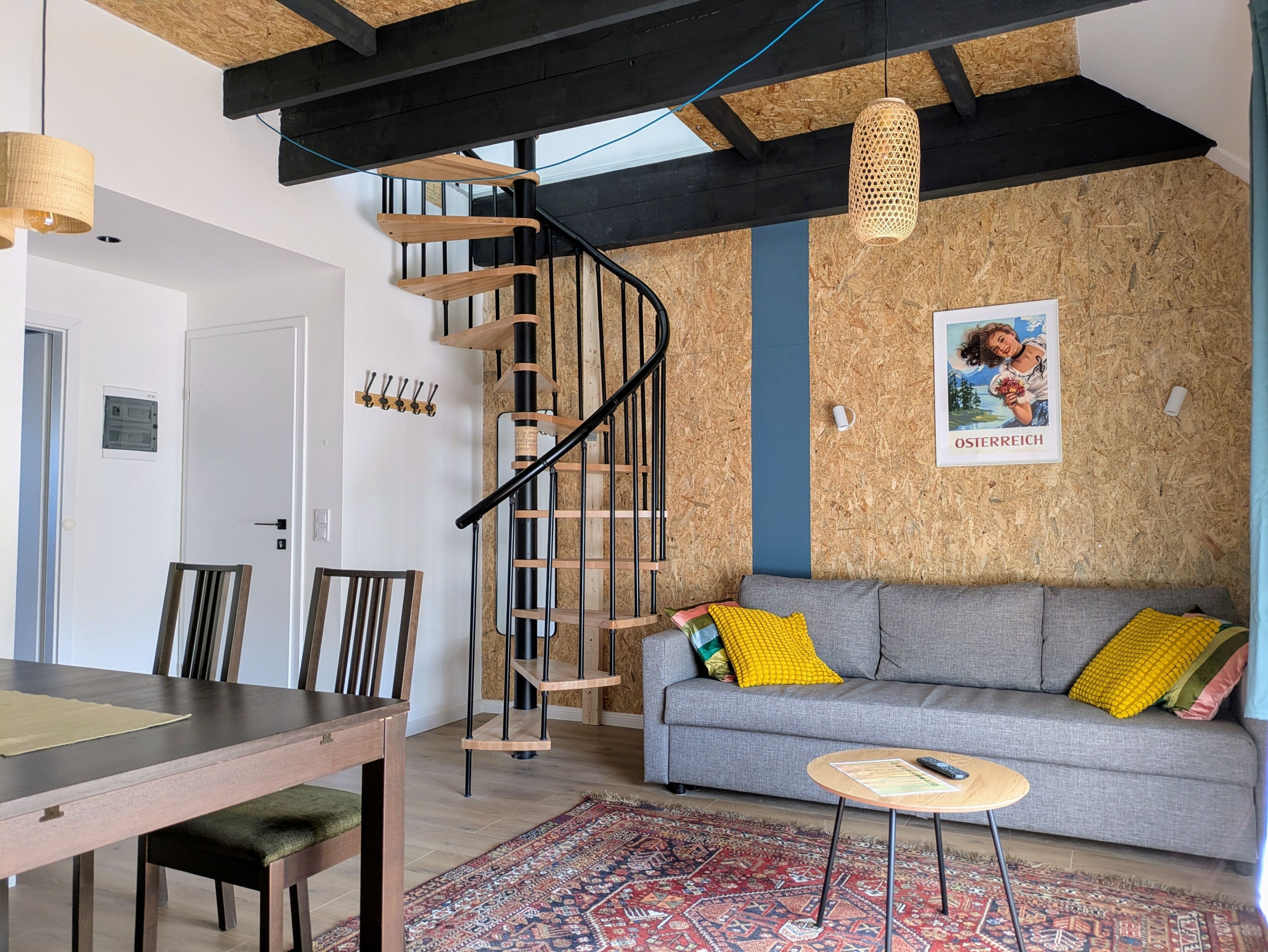 A modern living space with a wooden staircase, a grey sofa with colorful cushions, a dining table with chairs, and a patterned rug.