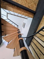 Una escalera de caracol con escalones de madera y barandillas metálicas, que conduce a una habitación con techo blanco y un cuadro en la pared.