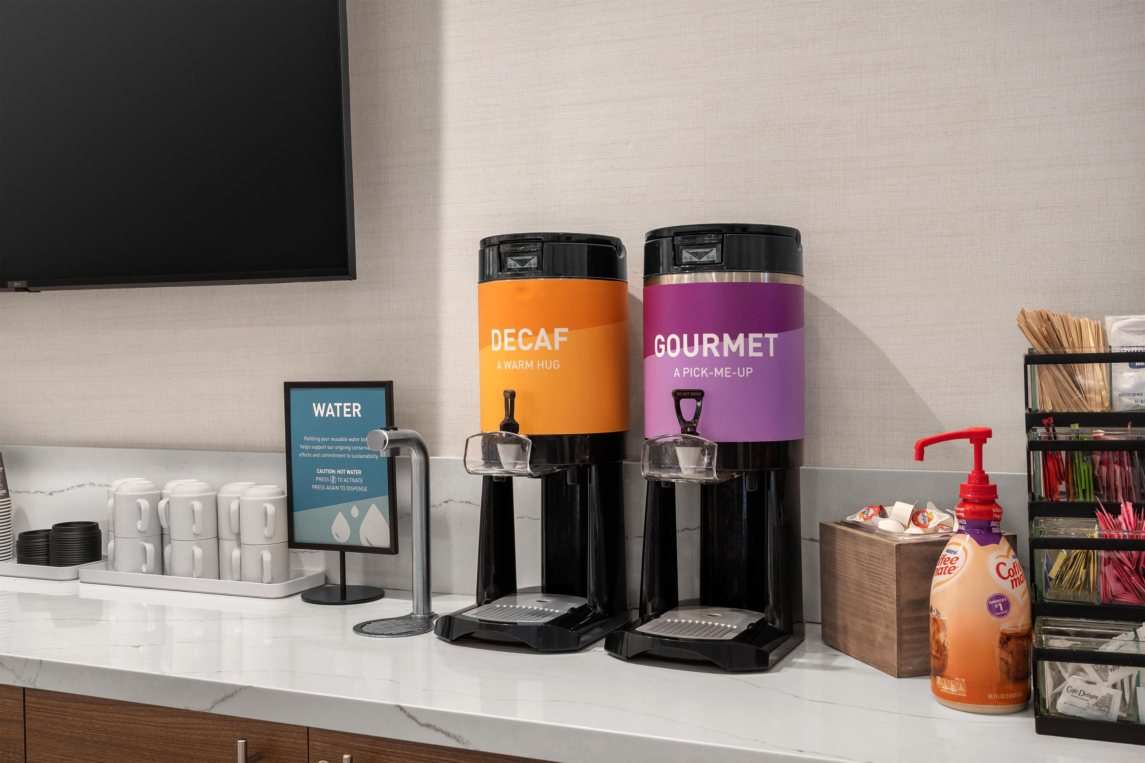 Una estación de dispensador de agua con dos dispensadores de bebidas, uno etiquetado 'DECAF' y otro 'GOURMET', una cafetera y una estación de café con un dispensador de leche.