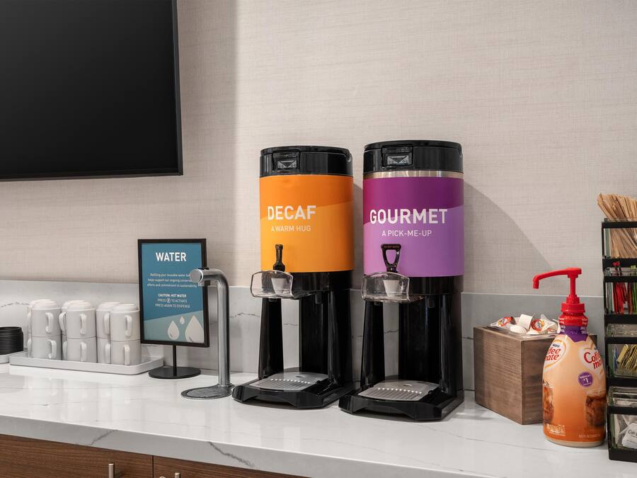 Una estación de dispensador de agua con dos dispensadores de bebidas, uno etiquetado 'DECAF' y otro 'GOURMET', una cafetera y una estación de café con un dispensador de leche.