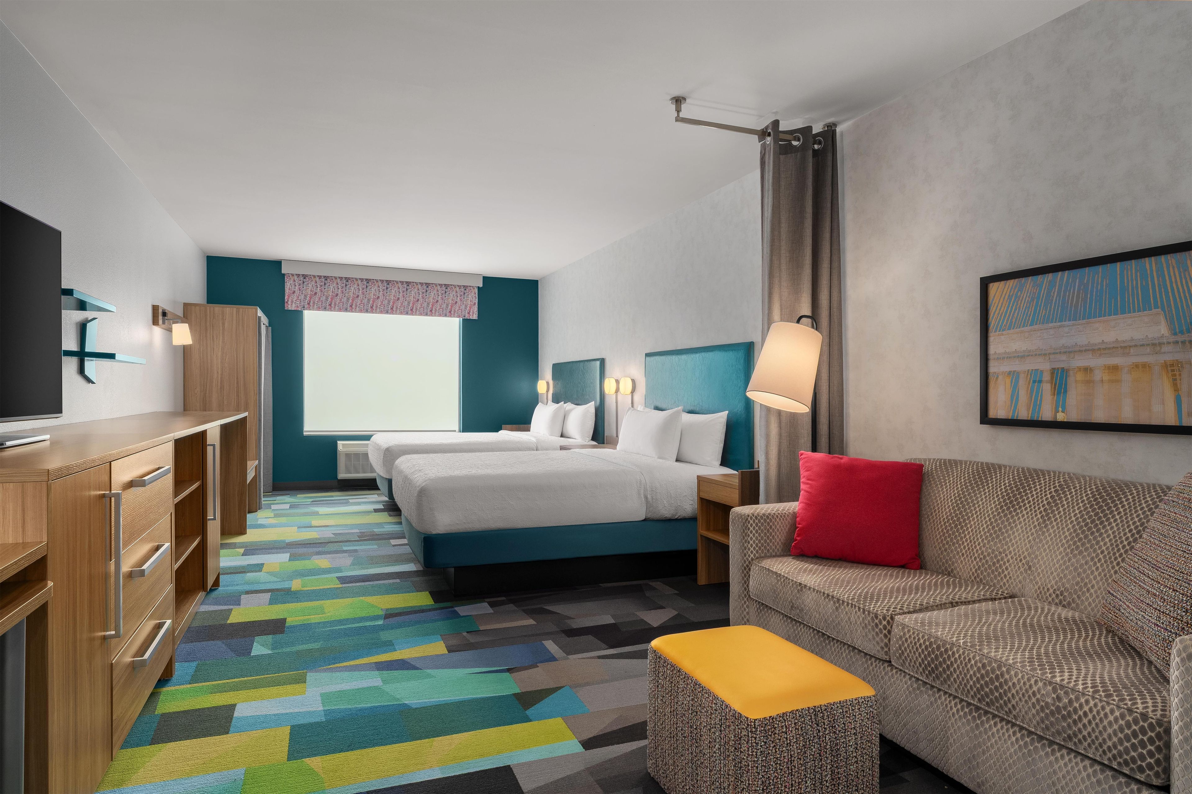 Quarto de hotel com cama, sofá, televisão, escrivaninha e tapete colorido.
