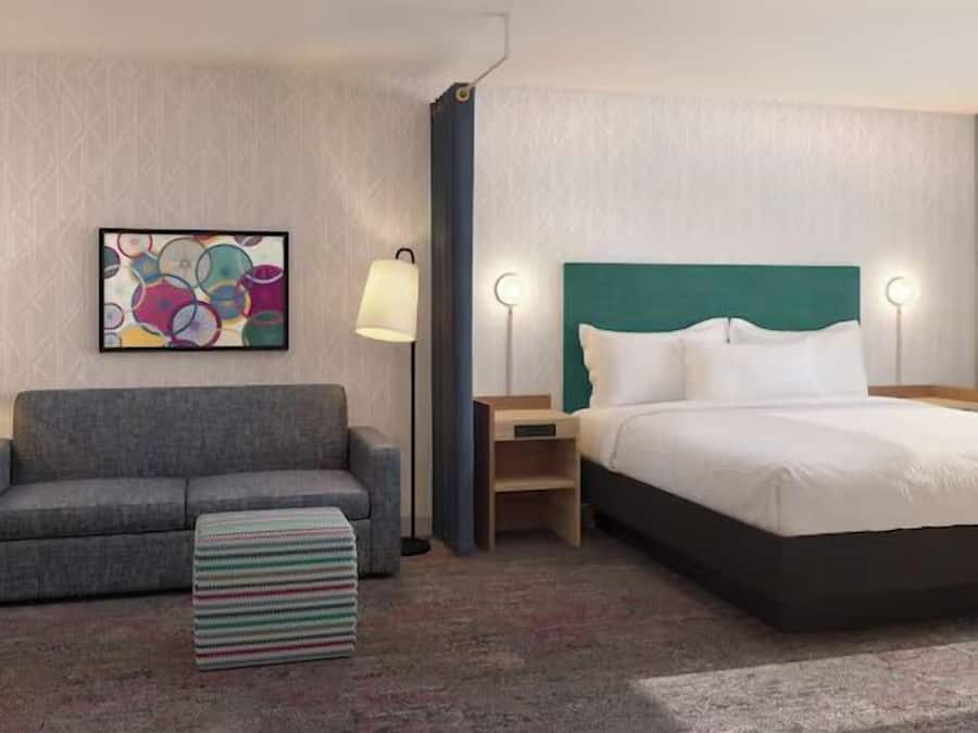 A hotel room with a gray sofa, a bed with a teal headboard, a colorful abstract painting, and a striped ottoman.