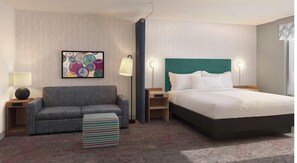 A hotel room with a gray sofa, a bed with a teal headboard, a colorful abstract painting, and a striped ottoman.