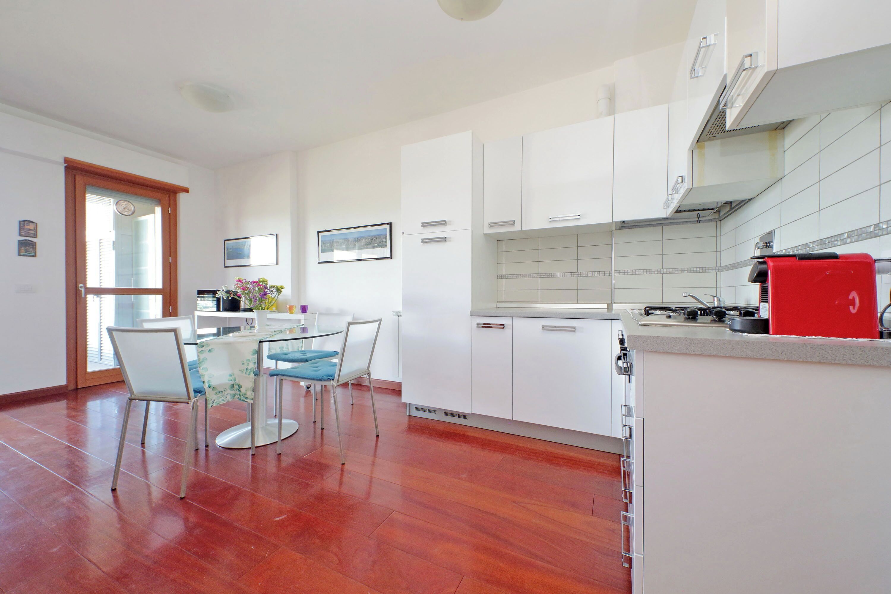 Une cuisine moderne avec des meubles blancs, un appareil électroménager rouge et un coin repas comprenant une table et des chaises en verre.