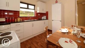 A compact kitchen with white cabinets, a red-tiled backsplash, and a dining area with a wooden table set for four. - Atlantic Coast (Ballyganny)