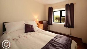 A bedroom with a bed, a bedside table, a lamp, a window with curtains, and a radiator. - Atlantic Coast (Ballyganny)
