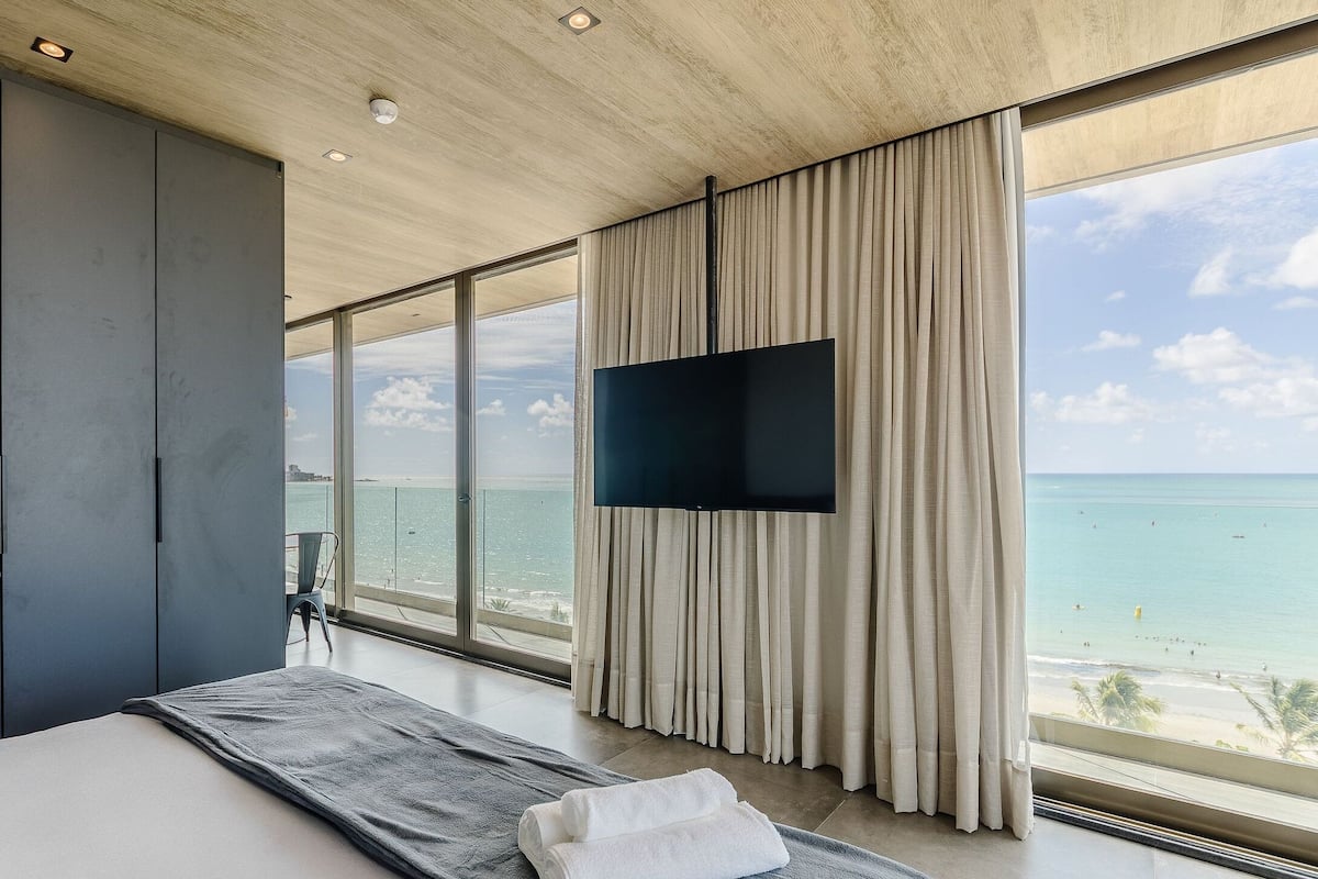 A modern bedroom with a large bed, a TV mounted on the wall, and a view of the beach.