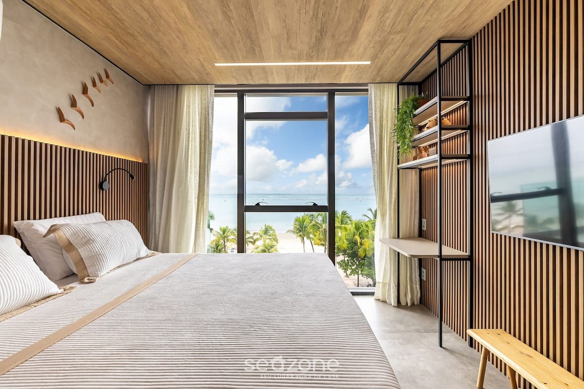 A modern bedroom with a large bed, a TV, and a view of the beach.
