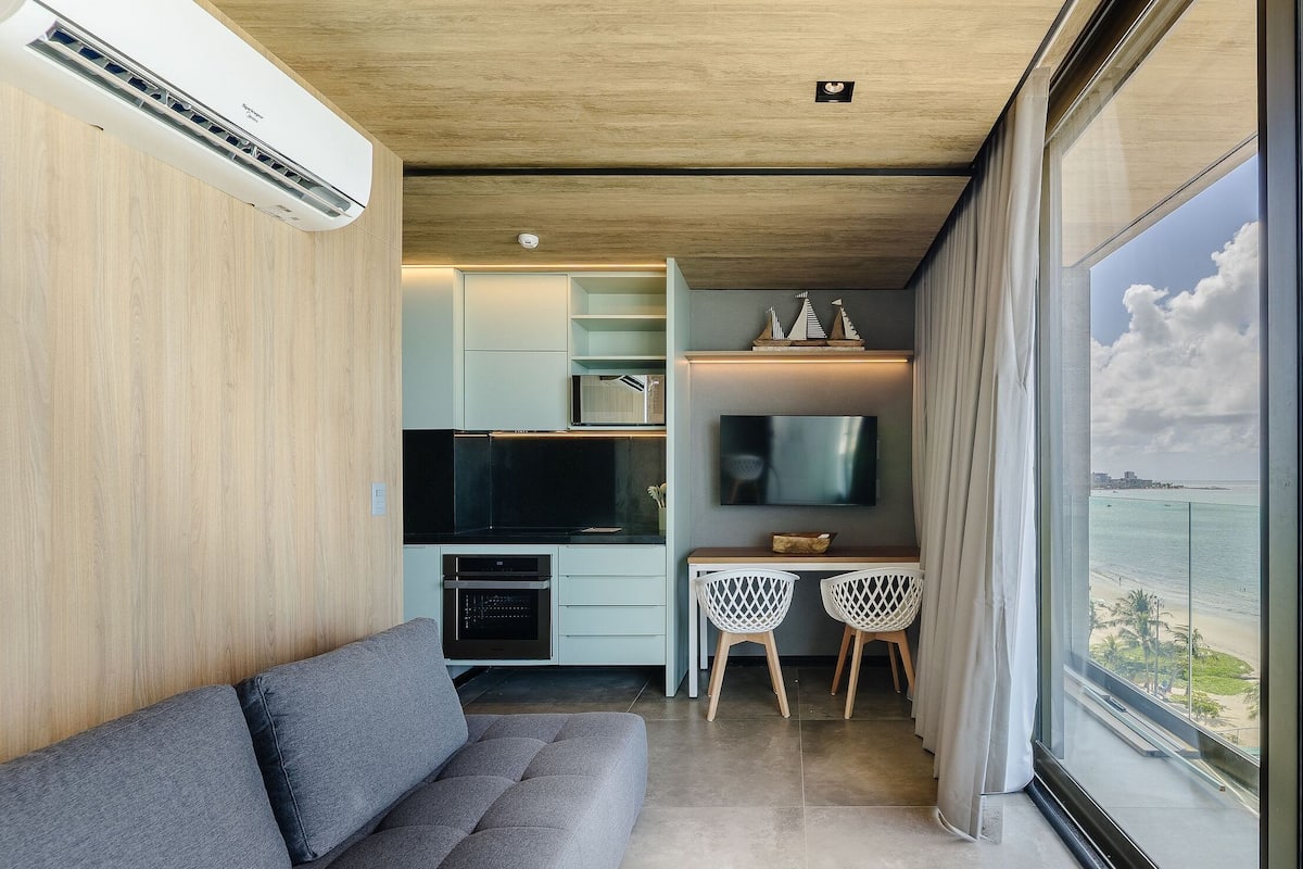 A modern living space with a kitchen, dining area, and a large window offering a view of the beach.