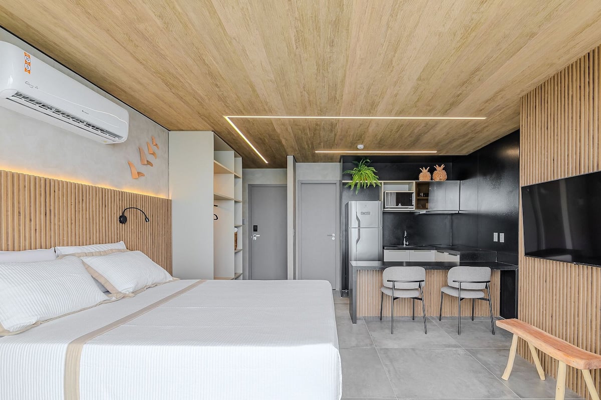 A modern hotel room with a bed, a small kitchenette, a TV, and a wooden ceiling.