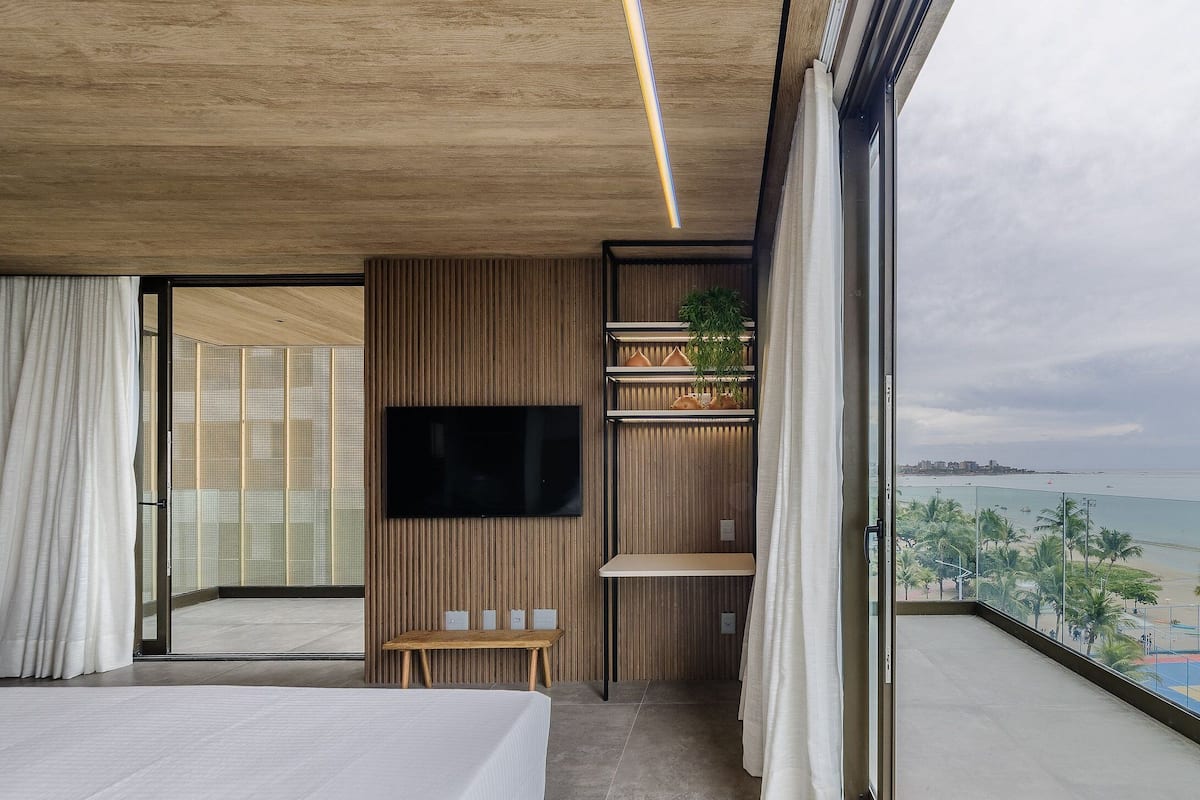 A modern bedroom with a large bed, a TV, a small table, and a balcony with a view of the beach.