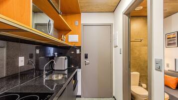 A compact kitchen with a black countertop, a sink, and a microwave. There is a door leading to a bathroom with a toilet and a shower.