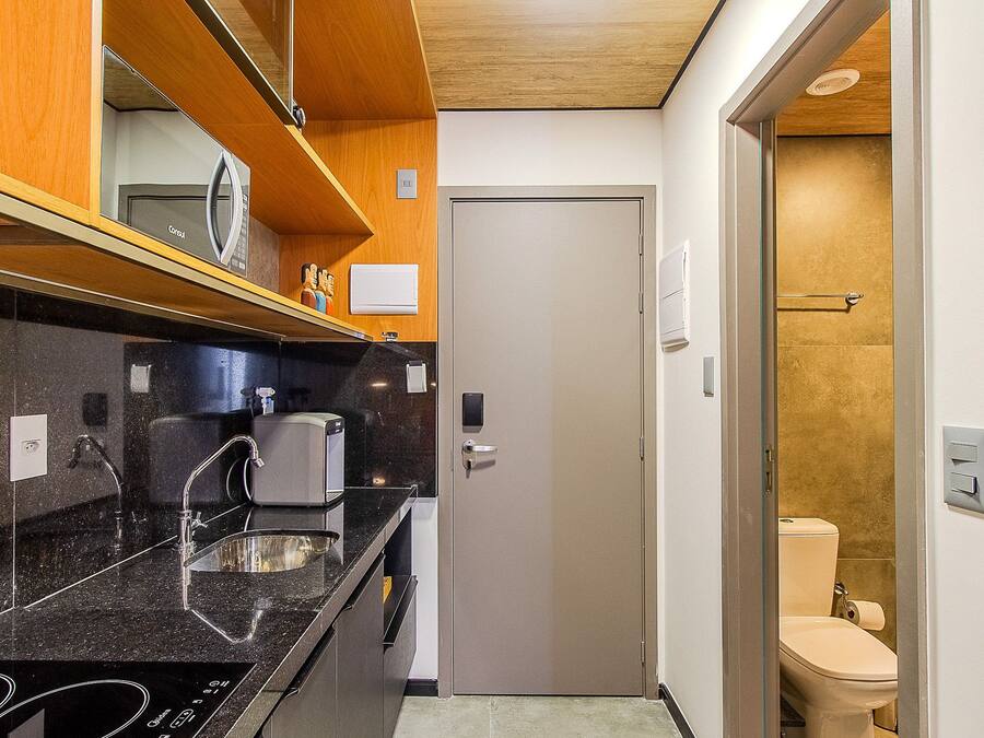 A compact kitchen with a black countertop, a sink, and a microwave. There is a door leading to a bathroom with a toilet and a shower.