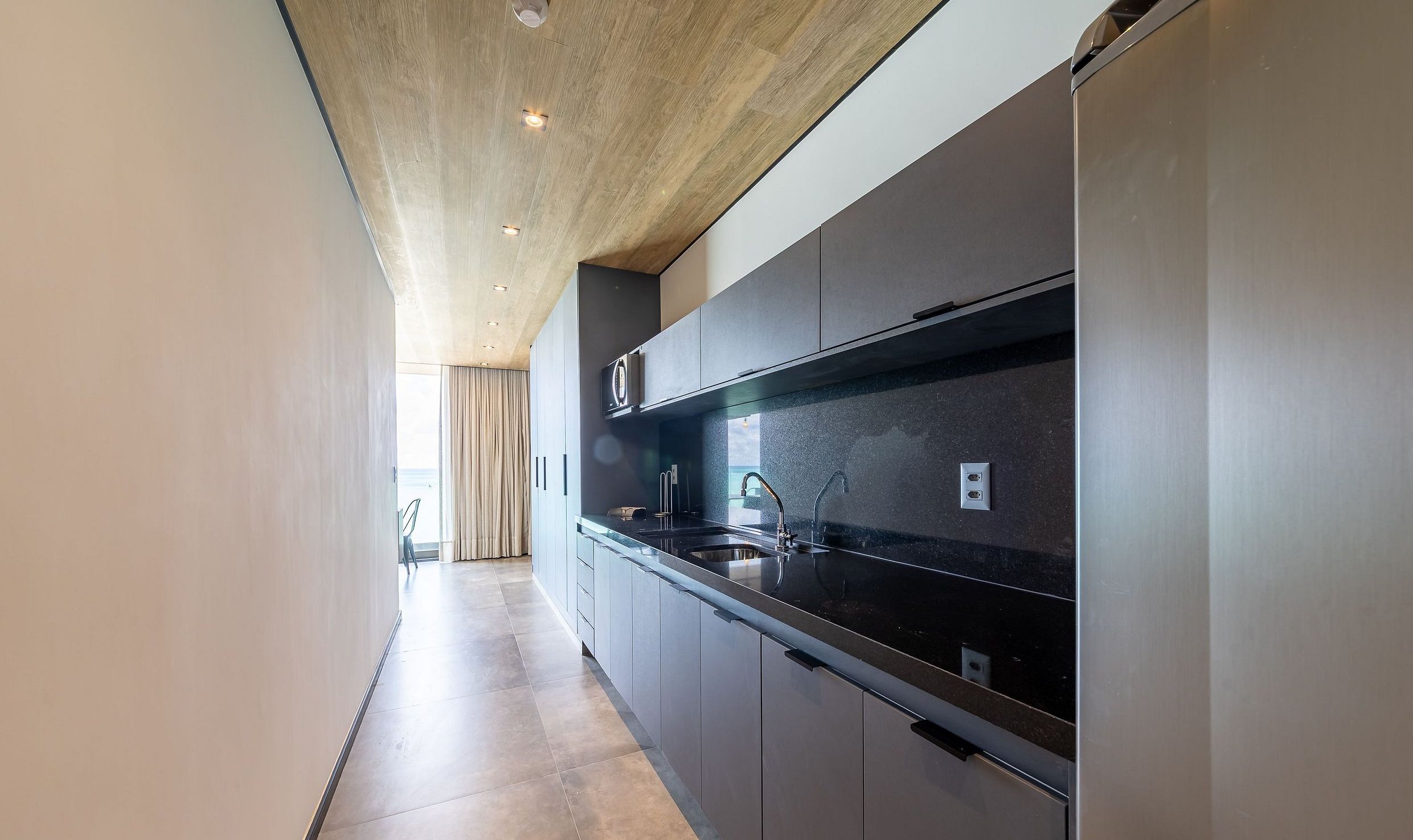 A modern kitchen with a long countertop, built-in appliances, and a wooden ceiling.