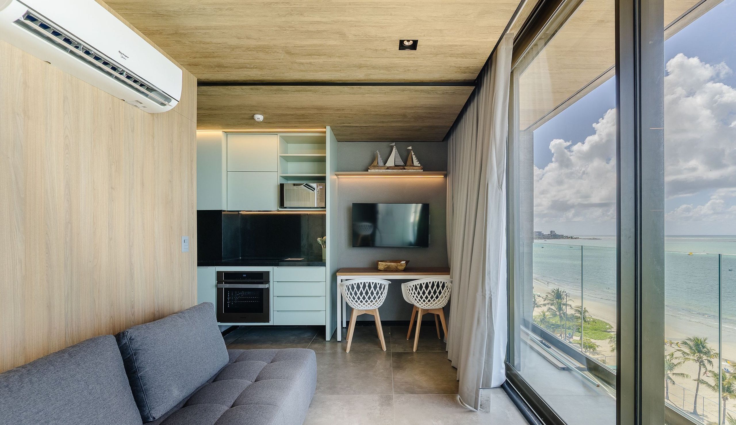 A modern living room with a sofa, a flat-screen TV, a dining area with two chairs, and a view of the ocean.
