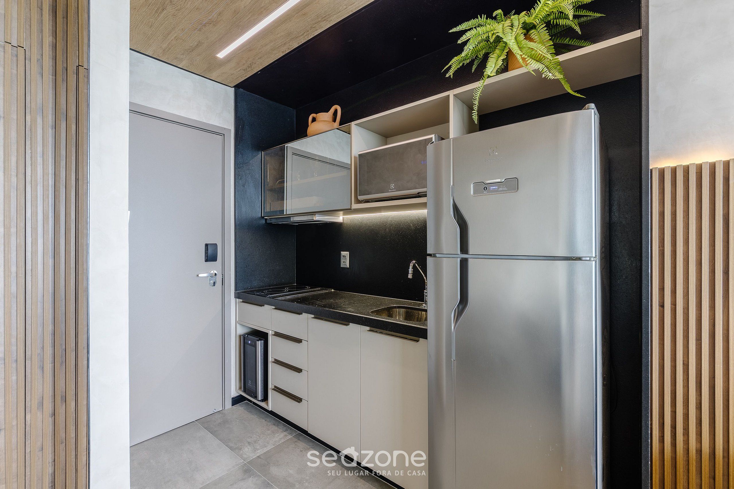 A modern kitchen with a stainless steel refrigerator, a built-in oven, and a microwave. There is a plant on the countertop and a wooden panel on the wall.