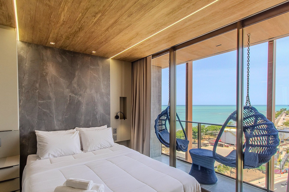 A bedroom with a large bed, a view of the ocean, and a hanging chair by the window.