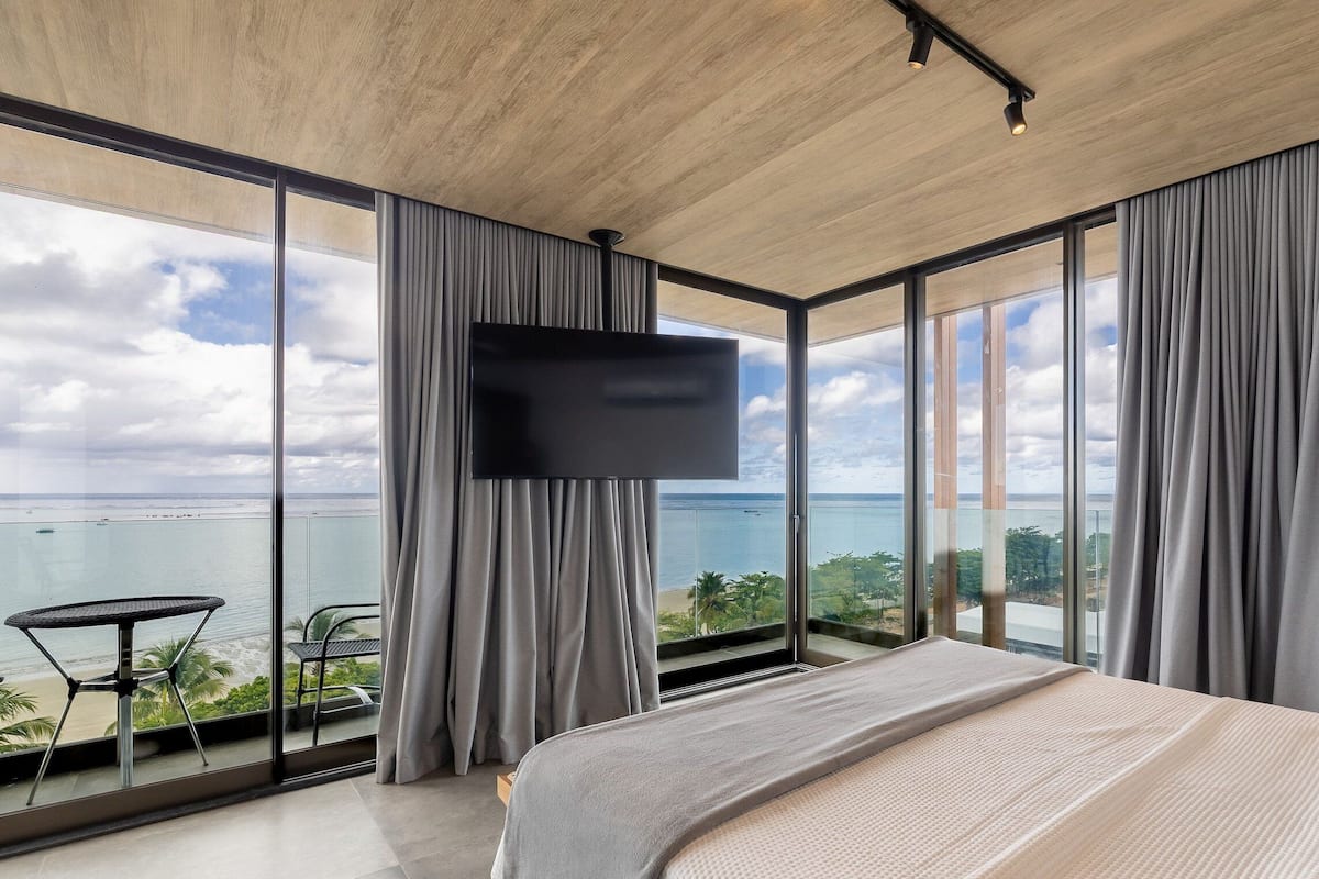 A modern bedroom with a large bed, a flat-screen TV, and a balcony with a view of the sea.