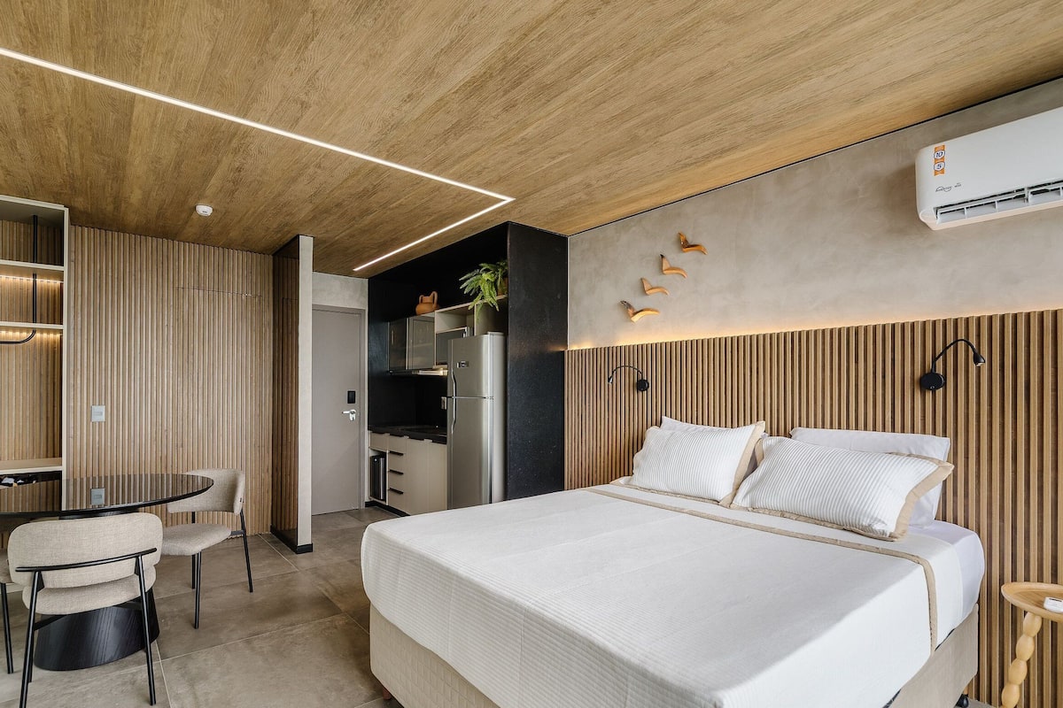 A modern hotel room with a wooden ceiling, a bed with white bedding, a dining area with a round table and chairs, and a kitchenette.