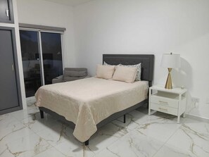 A bedroom with a bed, a nightstand, a lamp, a chair, and a sliding glass door. - Beautiful house with private pool, only 5 minutes from the sea, comfortable and safe. (Puerto Peñasco)