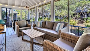 Un área de estar al aire libre con techo, amueblada con mobiliario de mimbre, una mesa de fútbol y amplias ventanas con vistas a un jardín exuberante.