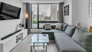 A modern living room with a large window, a flat-screen TV, a glass coffee table, and a grey sofa.