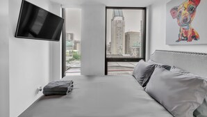 A modern hotel room with a bed, a flat-screen TV mounted on the wall, a large window with a city view, and a colorful painting on the wall.