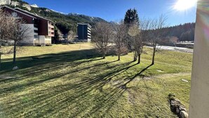 A grassy area with trees and buildings, including a modern structure and a tall building with a dark facade. - 1st floor, balcony, tv, 32m², Villard de Lans (Corrençon-en-Vercors)