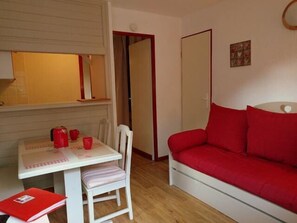 Um quarto pequeno e acolhedor com uma mesa branca, um sofá vermelho e uma cadeira. Há uma porta que dá para outro quarto e uma pequena estante com um livro.