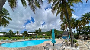 A swimming pool with lounge chairs and umbrellas, surrounded by palm trees and a mountainous backdrop. - Lovely Gentle Winds 2-bedroom condo! (St. Croix)