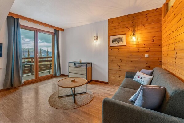 A cozy living room with a wooden wall, a sofa, a small table, and a view of a balcony. - Resort center, 3rd floor, fitness, swimming pool, sauna, balcony, tv, ski locker, 33m², Les Orres (Les Orres)