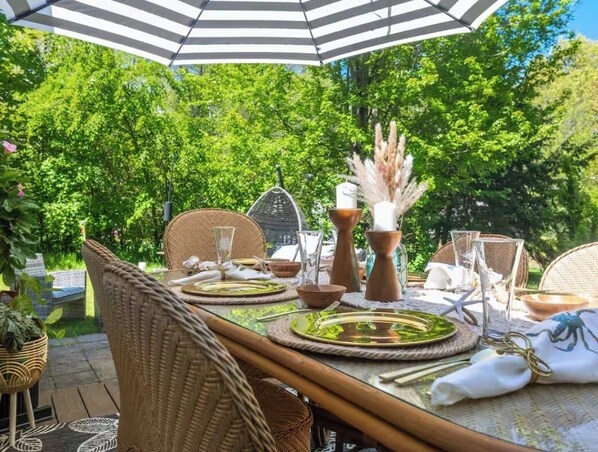 An outdoor dining area with wicker furniture, a striped umbrella, and a lush green backdrop.