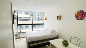 Ein modernes Schlafzimmer mit Bett, kleinem Tisch und Stuhl. Ein Fenster bietet Blick auf Gebäude.