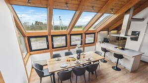 Un espace repas avec une table et des chaises en bois, une cuisine dotée d’un plan de travail blanc et de grandes fenêtres offrant une vue sur l’extérieur.