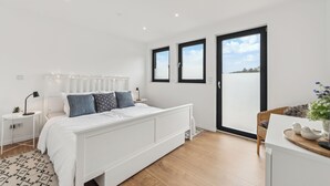 Une chambre moderne avec un lit blanc, une petite table de chevet, un fauteuil en osier et une vue sur l’extérieur grâce à une grande fenêtre.