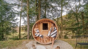 A wooden sauna in a forest setting with a bench and a chair nearby.