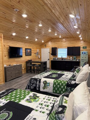 Een kamer in chaletstijl met houten wanden en plafond, twee bedden met groen en witte beddengoed, een bureau met stoelen, een televisie en een kitchenette.