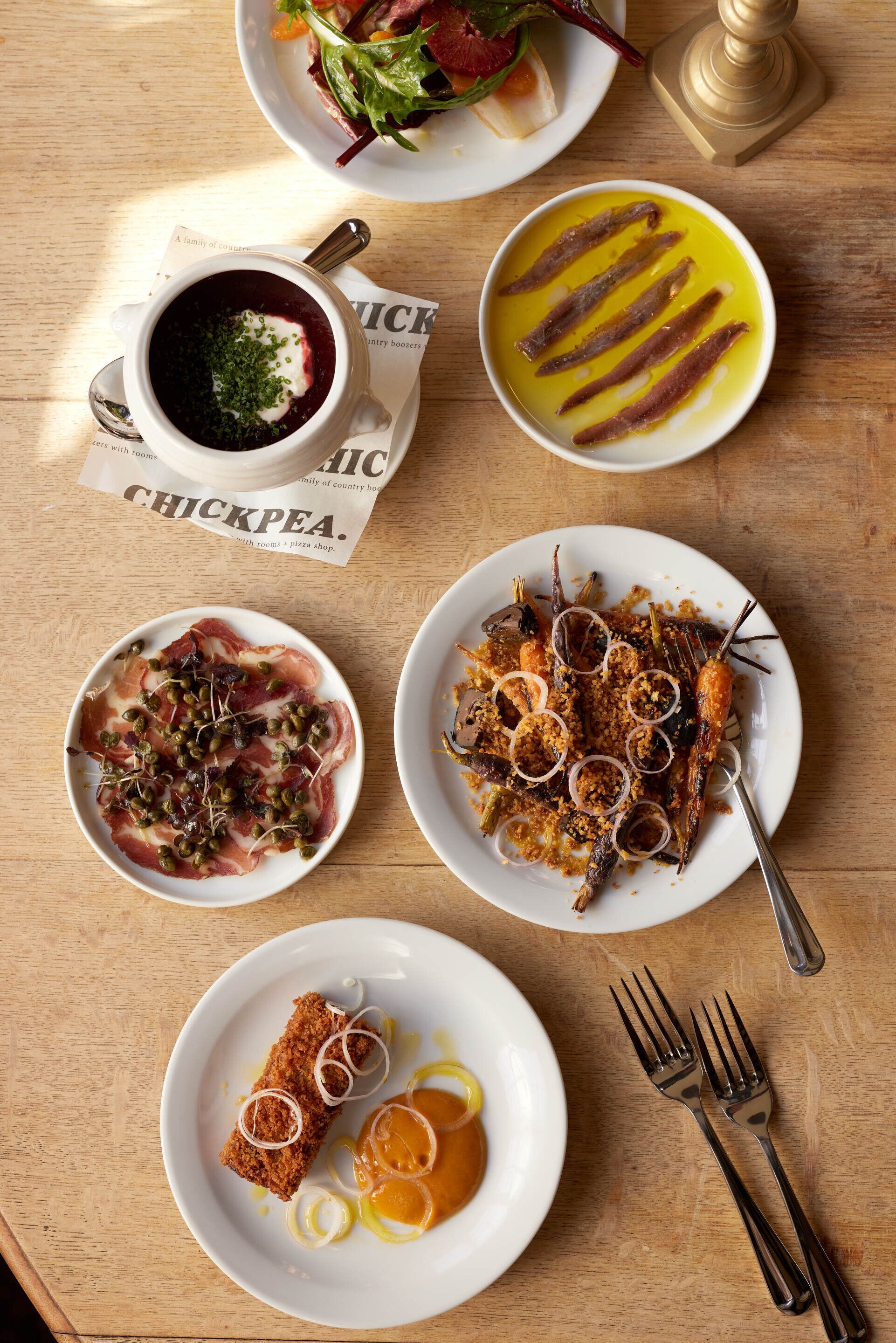 A wooden table with various dishes including a bowl of soup, a plate with meat and olives, and a plate with a fried item.