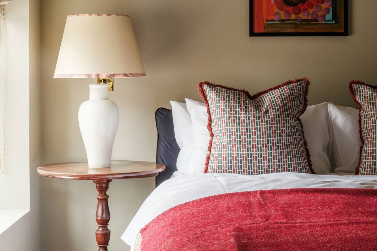 Una camera d'albergo con un letto, un comodino con una lampada, una coperta decorata e un quadro appeso al muro.