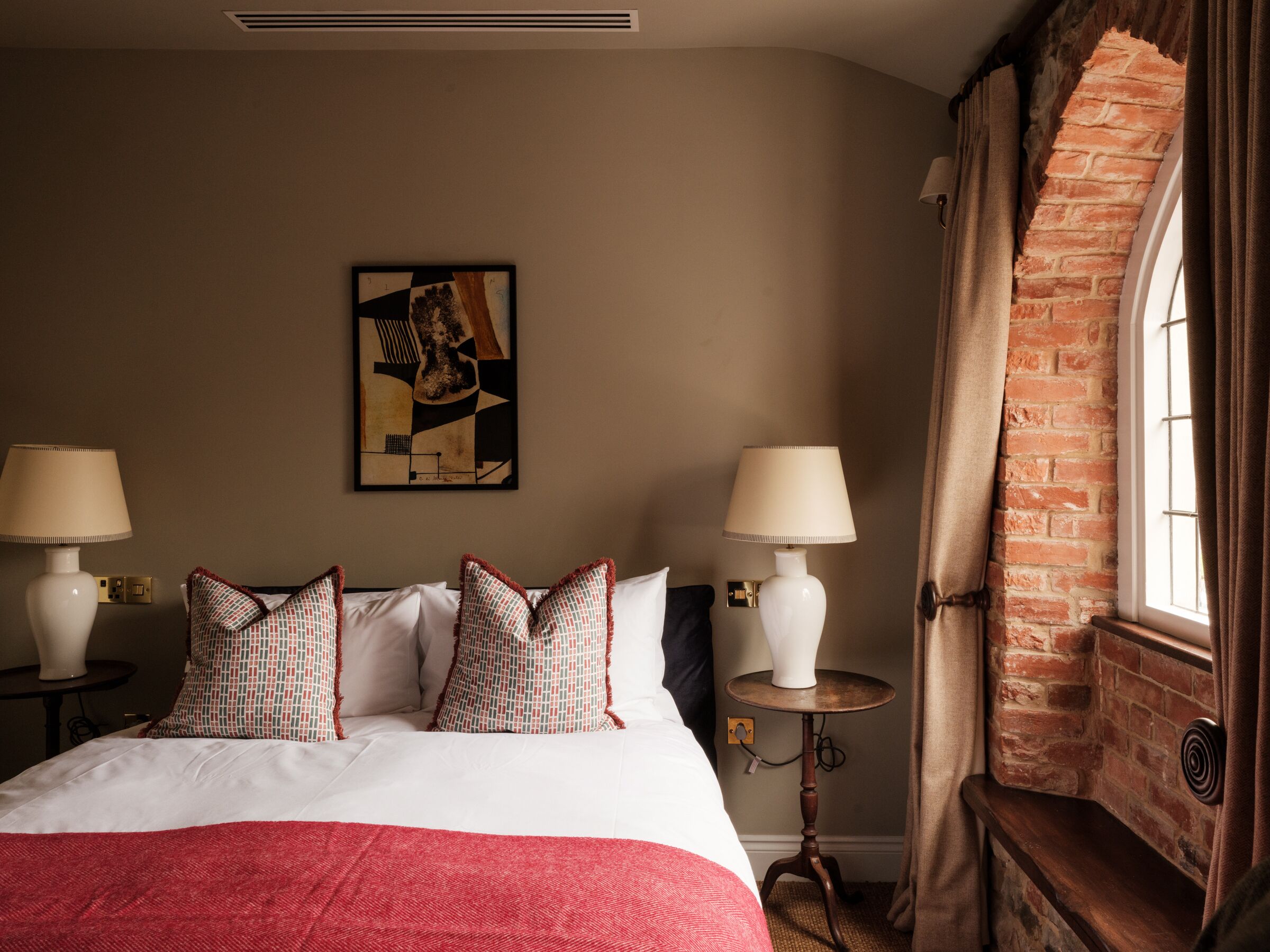 A hotel room with a large bed, two bedside lamps, a small table, a brick wall, and a window with curtains.