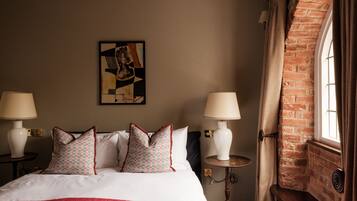 A hotel room with a large bed, two bedside lamps, a small table, a brick wall, and a window with curtains.