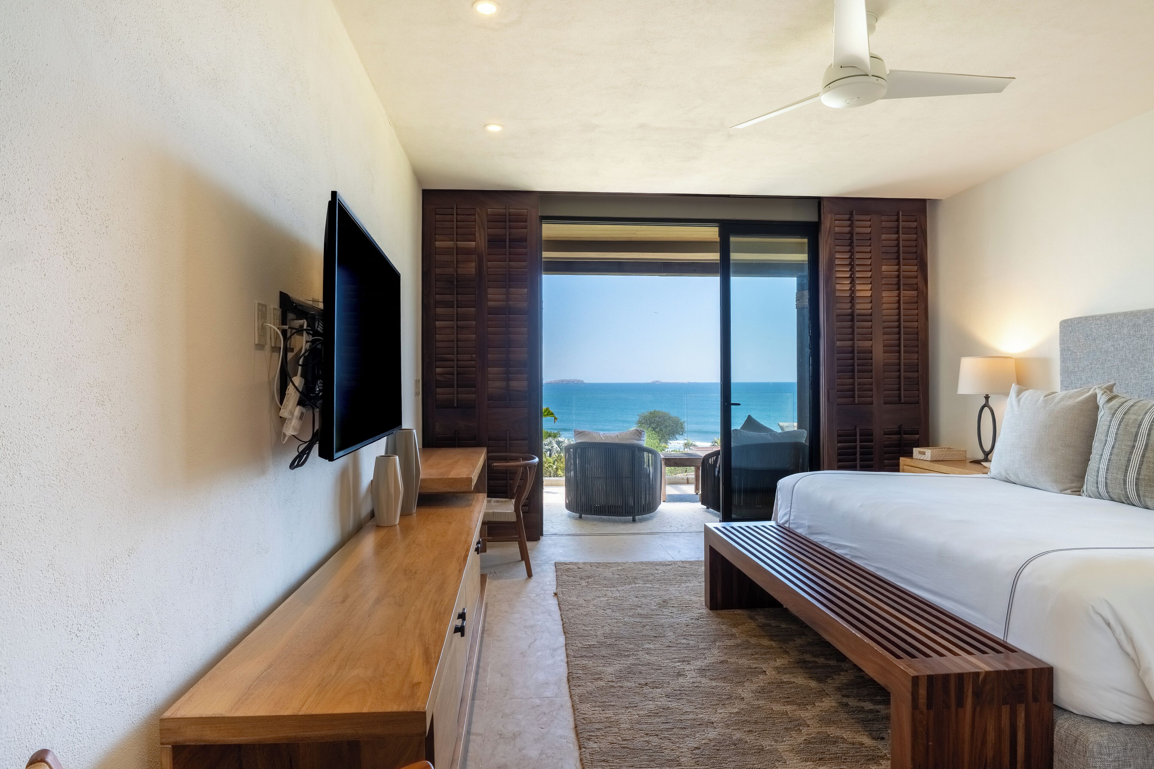 A modern hotel room with a large bed, a desk, a television, and a view of the ocean.