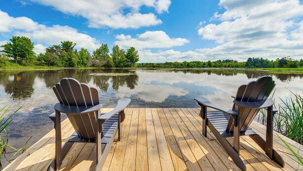 A wooden deck with two Adirondack chairs facing a calm lake, surrounded by trees and a clear sky. - Riverfront Retreat/Hot Tub/ Short Walk to Downtown (Bellaire)