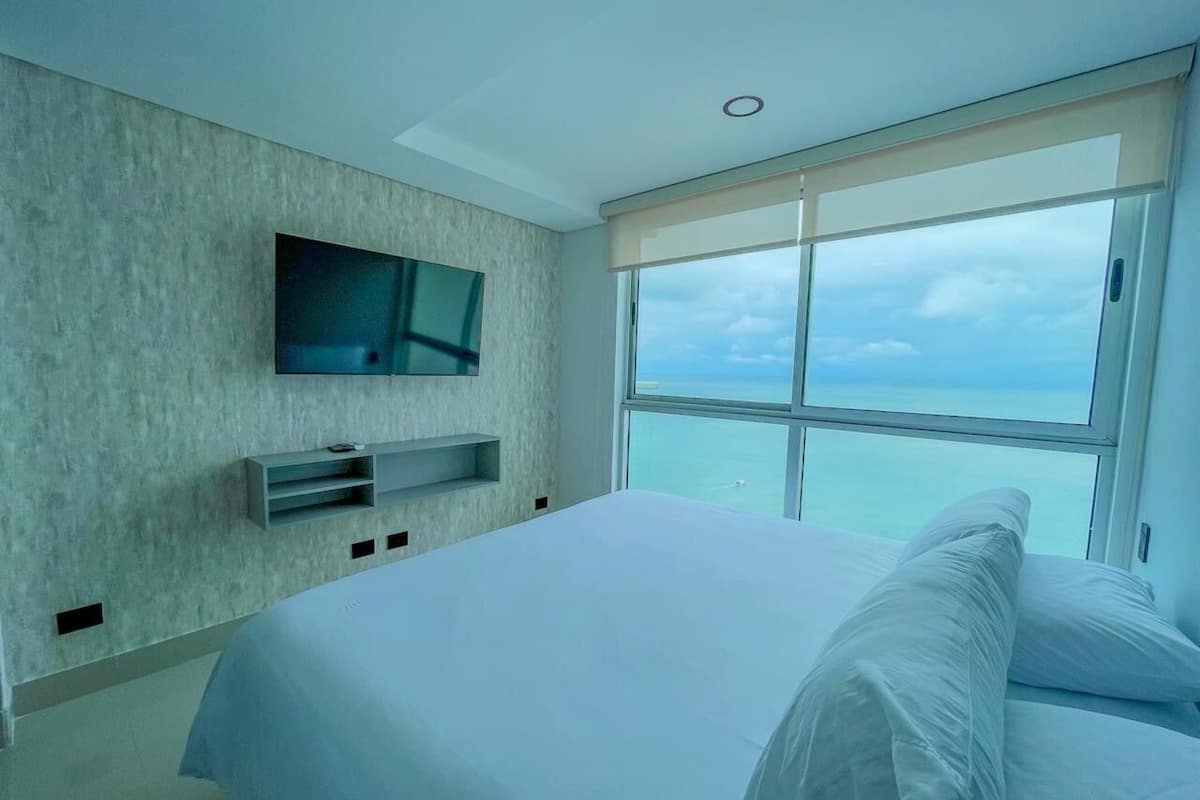 A modern bedroom with a large bed, a mounted TV, and a view of the ocean.