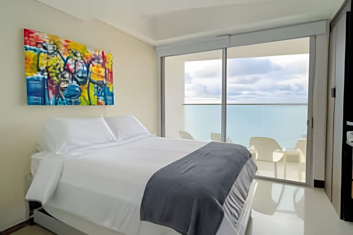 A modern bedroom with a large bed, a colorful abstract painting, and a balcony with a view of the sea.