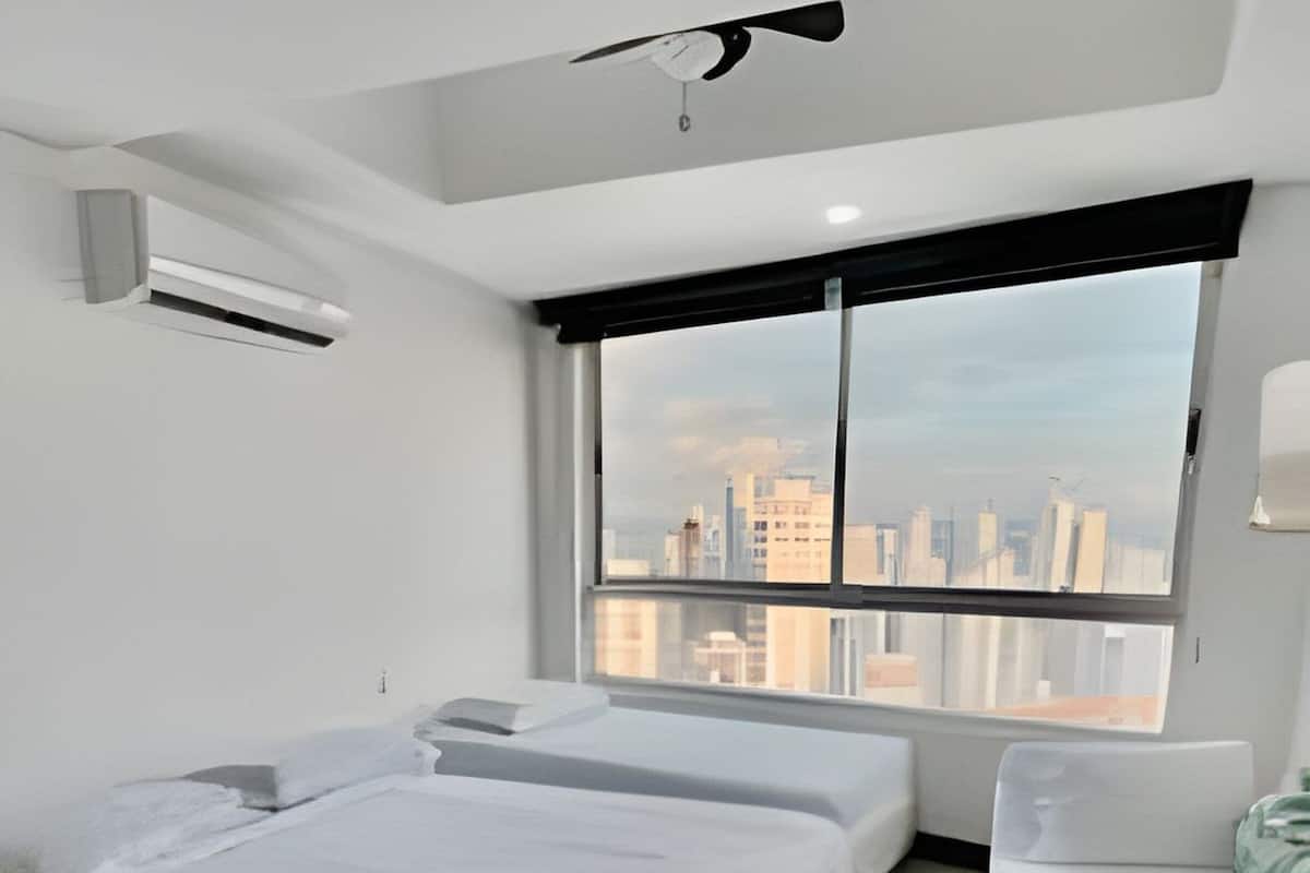 A modern hotel room with two beds, a large window with a city view, and a ceiling fan.