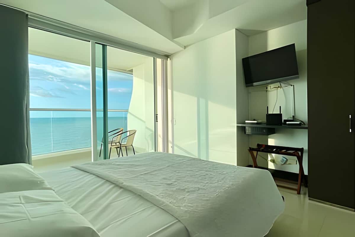A hotel room with a bed, a TV, a desk with a chair, and a balcony with a view of the sea.