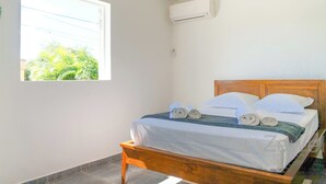 Et soveværelse med en træseng, hvide sengetøj, et vindue med udsigt til grøn natur, og en aircondition-enhed på væggen.