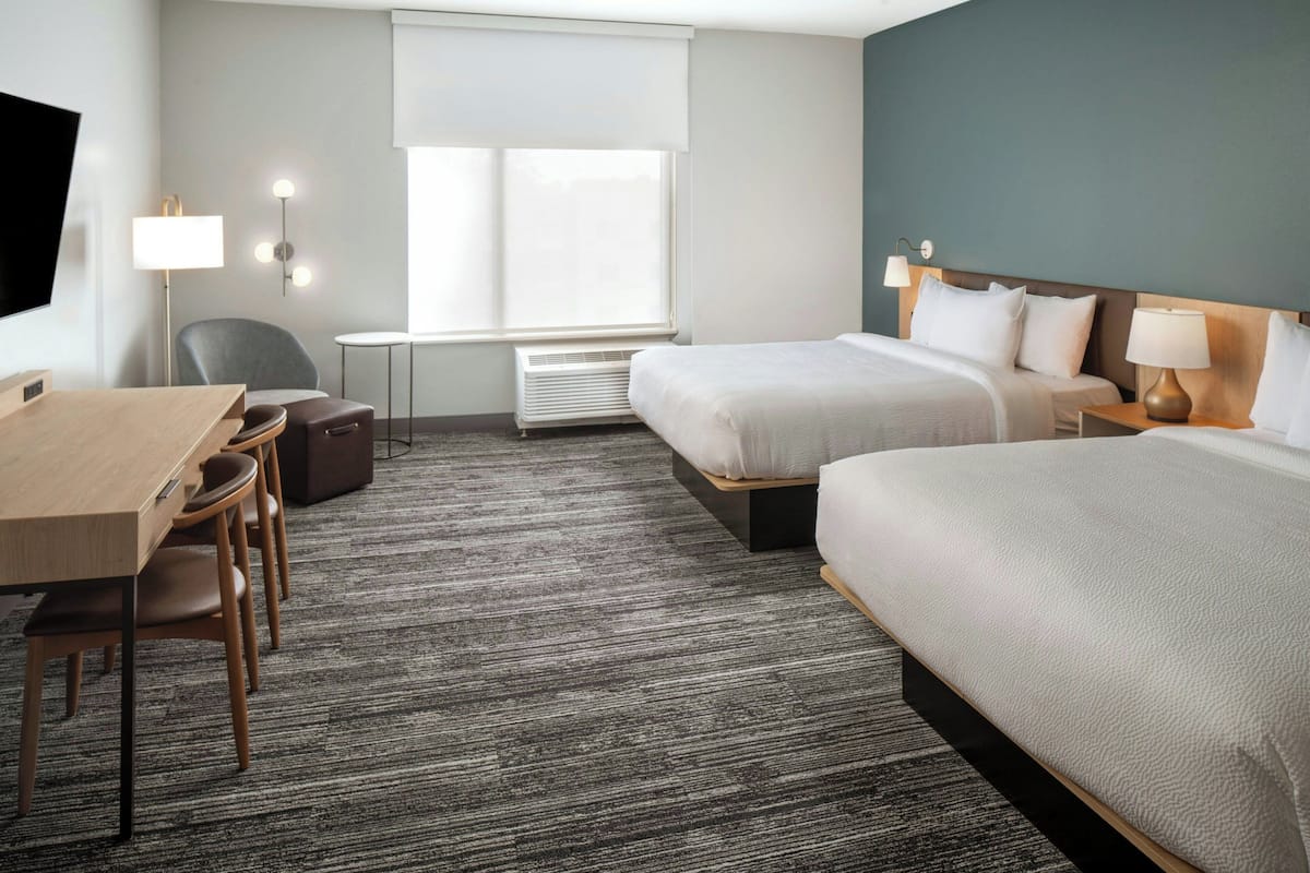 Ein Hotelzimmer mit zwei Betten, einem Schreibtisch mit Stühlen, einem Fernseher und einem Fenster mit Jalousien.
